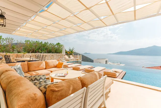 A shaded terrace with deep sofas and cushions beside an infinity pool, looking across the water to distant hills
