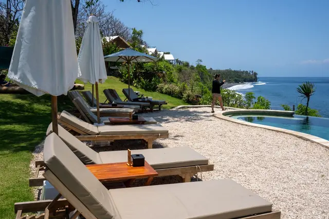 Cushioned sun loungers and white parasols on a lawn above a small pool, with the coastline beyond