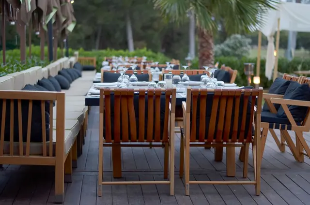 An outdoor dining terrace with timber chairs, laid tables and cushioned banquettes under the trees at dusk