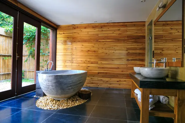 A stone bathtub on a bed of pebbles in a timber-panelled bathroom with glass doors to a garden