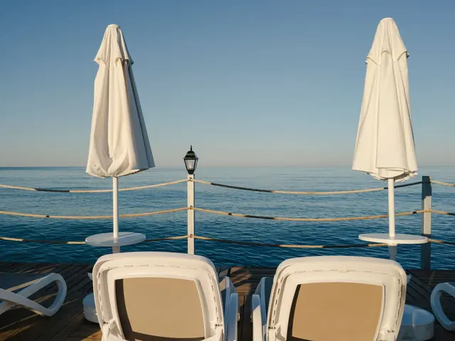 Two white loungers and furled parasols on a seaside deck facing calm blue water