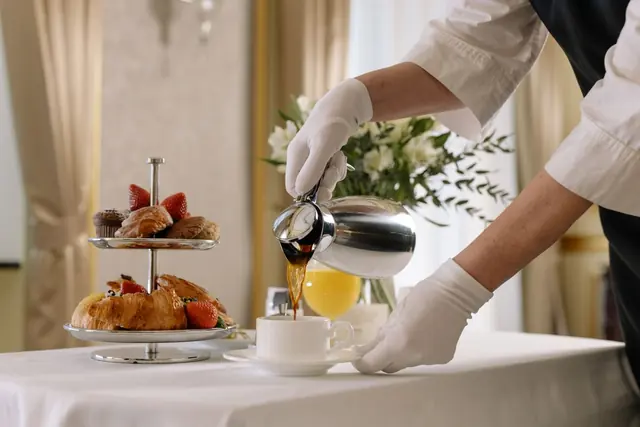A gloved server pouring coffee beside a tiered stand of pastries and strawberries on a white tablecloth