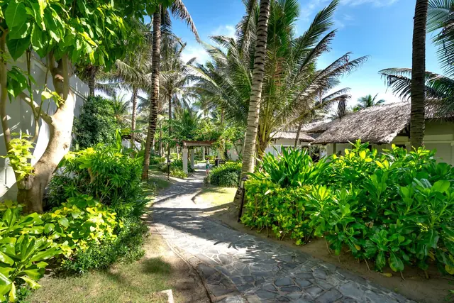 A stone path winding through palms and flowering shrubs towards thatched garden cottages