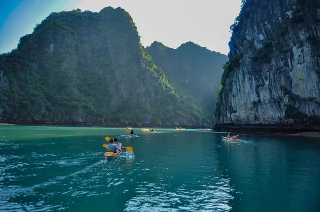 Kayakers paddling across a teal bay beneath tall forested limestone cliffs
