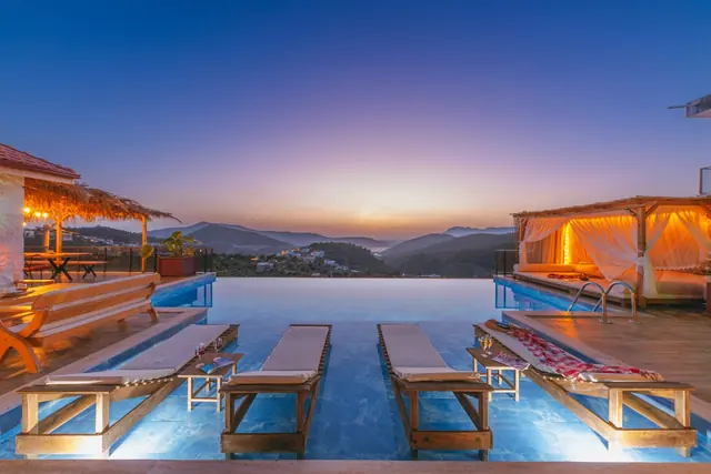Sun loungers set into the shallow edge of an infinity pool at dusk, with lit cabanas and hills beyond