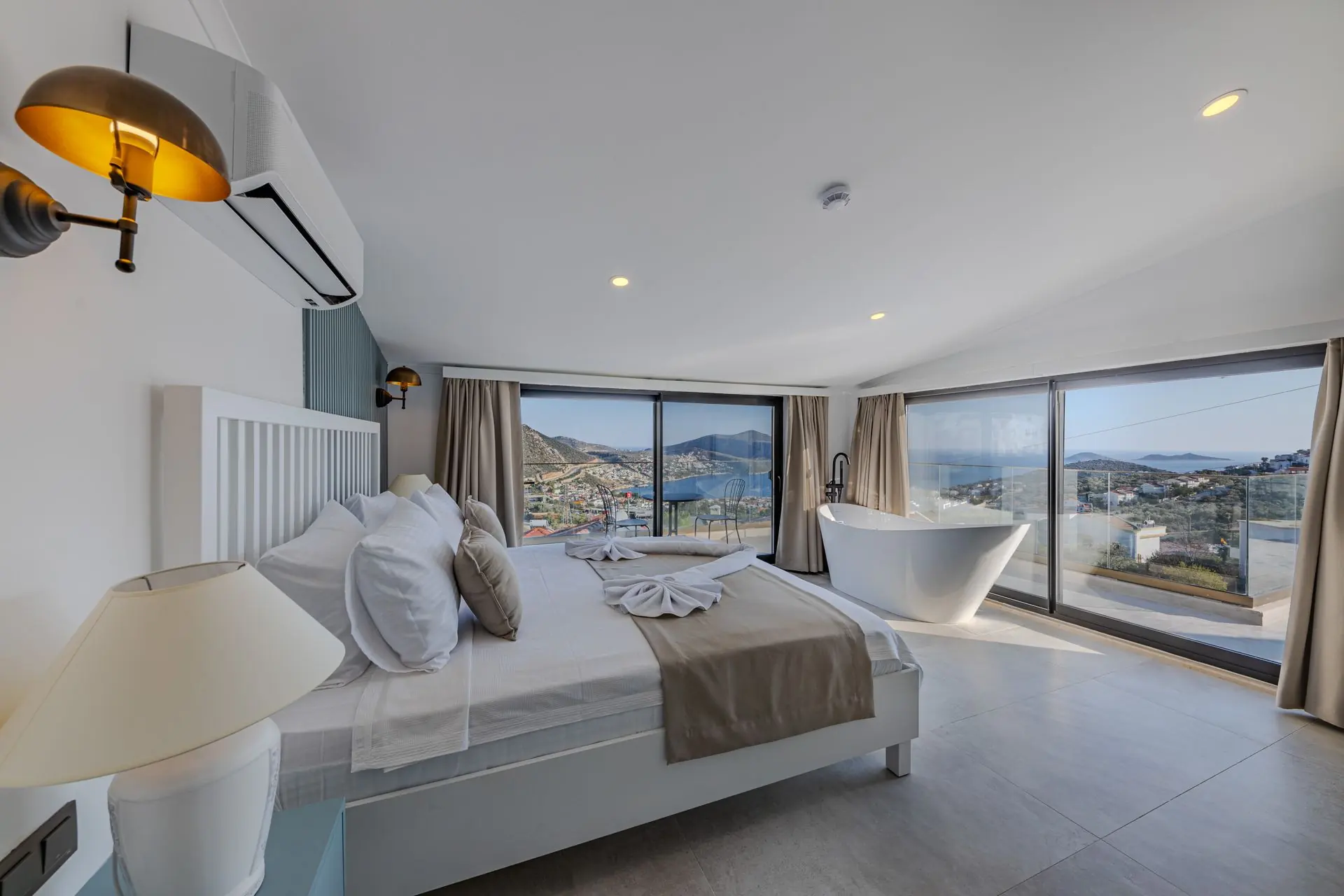 A bright bedroom with a made-up bed, a freestanding bath and floor-to-ceiling windows looking over the sea