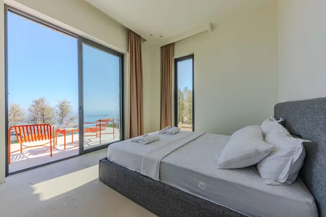 A light, minimal bedroom with a grey upholstered bed and sliding glass doors to a balcony above the sea
