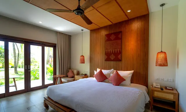 A calm bedroom with a timber feature wall, a large bed with coral cushions and glass doors opening to a garden