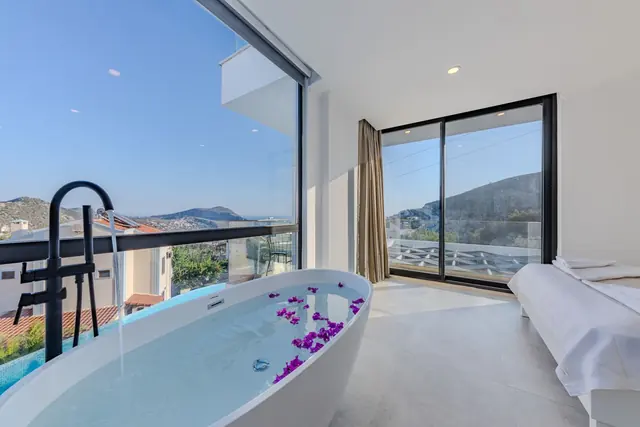 A freestanding bath scattered with petals beside a corner window looking over hills and the sea