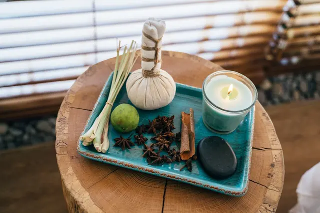 Herbal compresses, a lit candle, lime and cinnamon sticks arranged on a teal tray on a wooden stool
