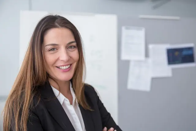 Portrait of a smiling professional in a dark blazer, arms folded, in a bright office