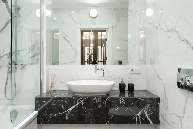 A white marble bathroom with a black stone vanity, round basin and glass shower