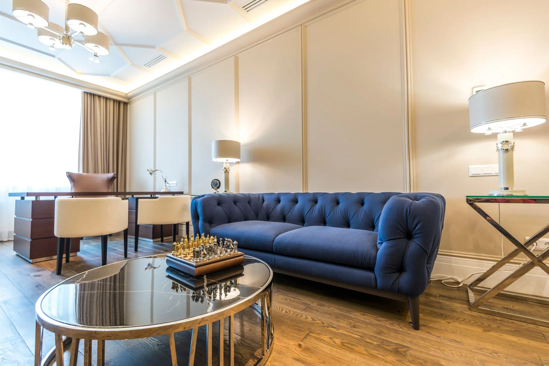 A bright living room with a navy tufted sofa, brass side tables and tall curtained windows