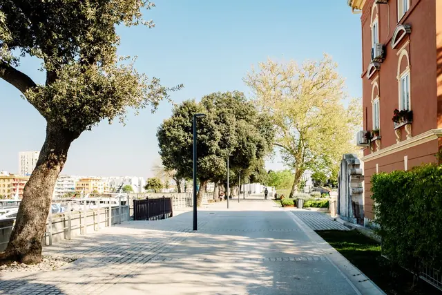 A sunny paved walkway shaded by mature trees beside a terracotta-coloured residential building