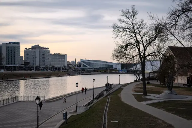 A riverside promenade with street lamps and a lawn, and apartment blocks across the water at dusk