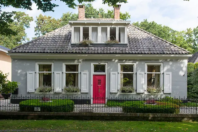 A pale grey cottage with white shutters, a red front door and a clipped front garden behind railings