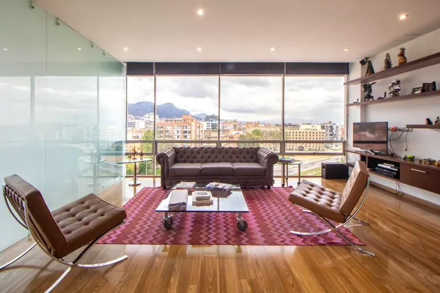 A penthouse living room with floor-to-ceiling windows, leather chairs and a view over the city to distant hills