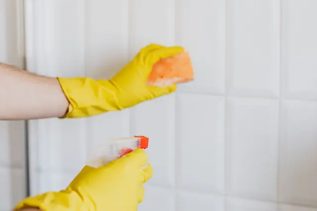 Gloved hands scrubbing white bathroom tiles with a sponge