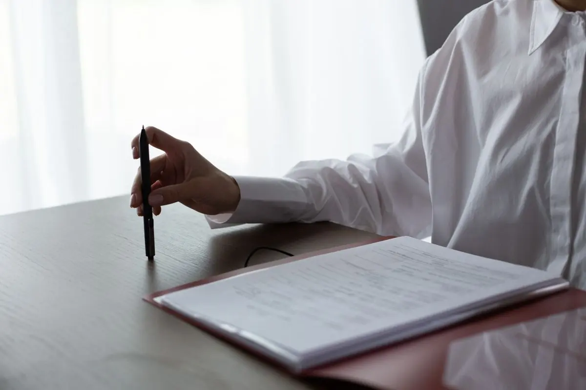 A person in a white shirt holding a pen over a folder of printed documents on a dark desk