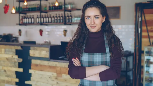 A café owner in a checked apron standing with folded arms in front of a wooden counter