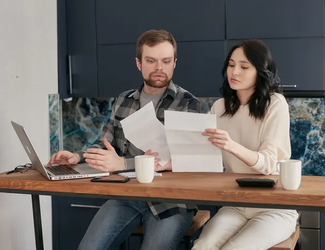 Two people at a kitchen table reading printed documents together, with a laptop and mugs nearby