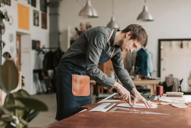 A craftsperson in a canvas apron leaning over a workbench to cut leather in a bright workshop