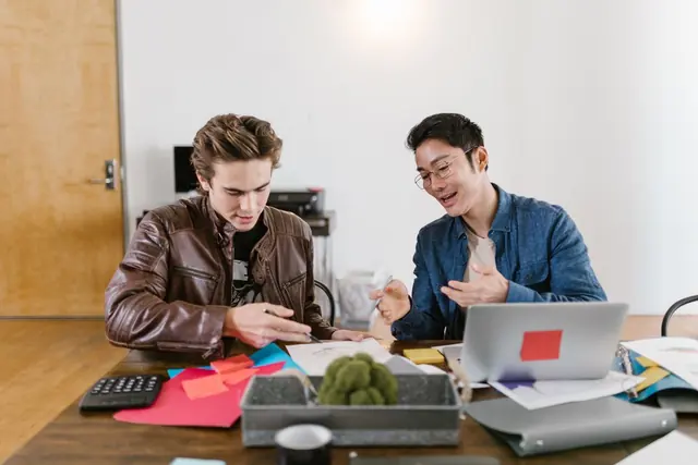 Two people at a shared table with a laptop, sticky notes and papers, talking through a plan