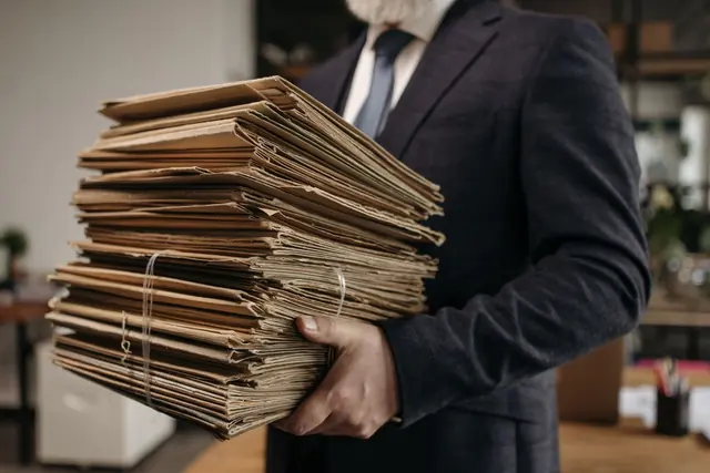 A person in a suit carrying a tall stack of brown card folders tied with string
