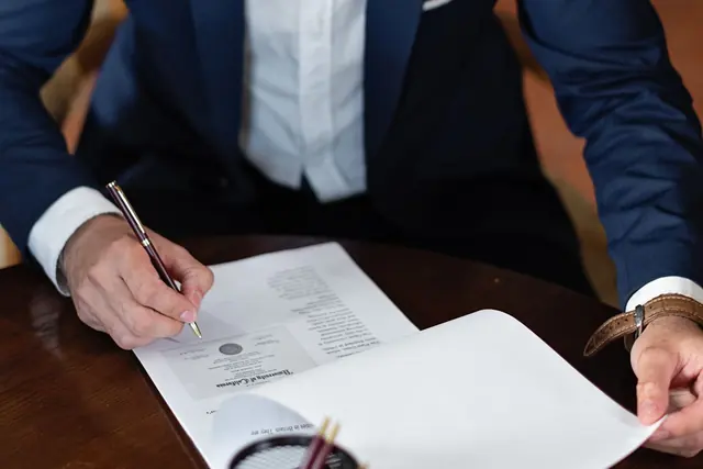 A person in a navy suit signing a printed document at a dark wooden desk
