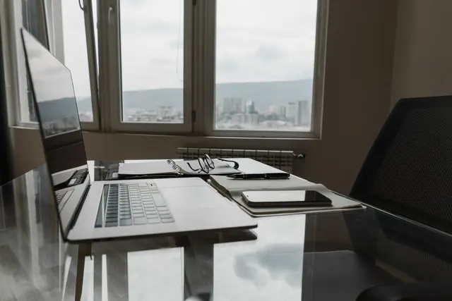 A glass desk by a large window with a laptop, a notebook and reading glasses, overlooking a city