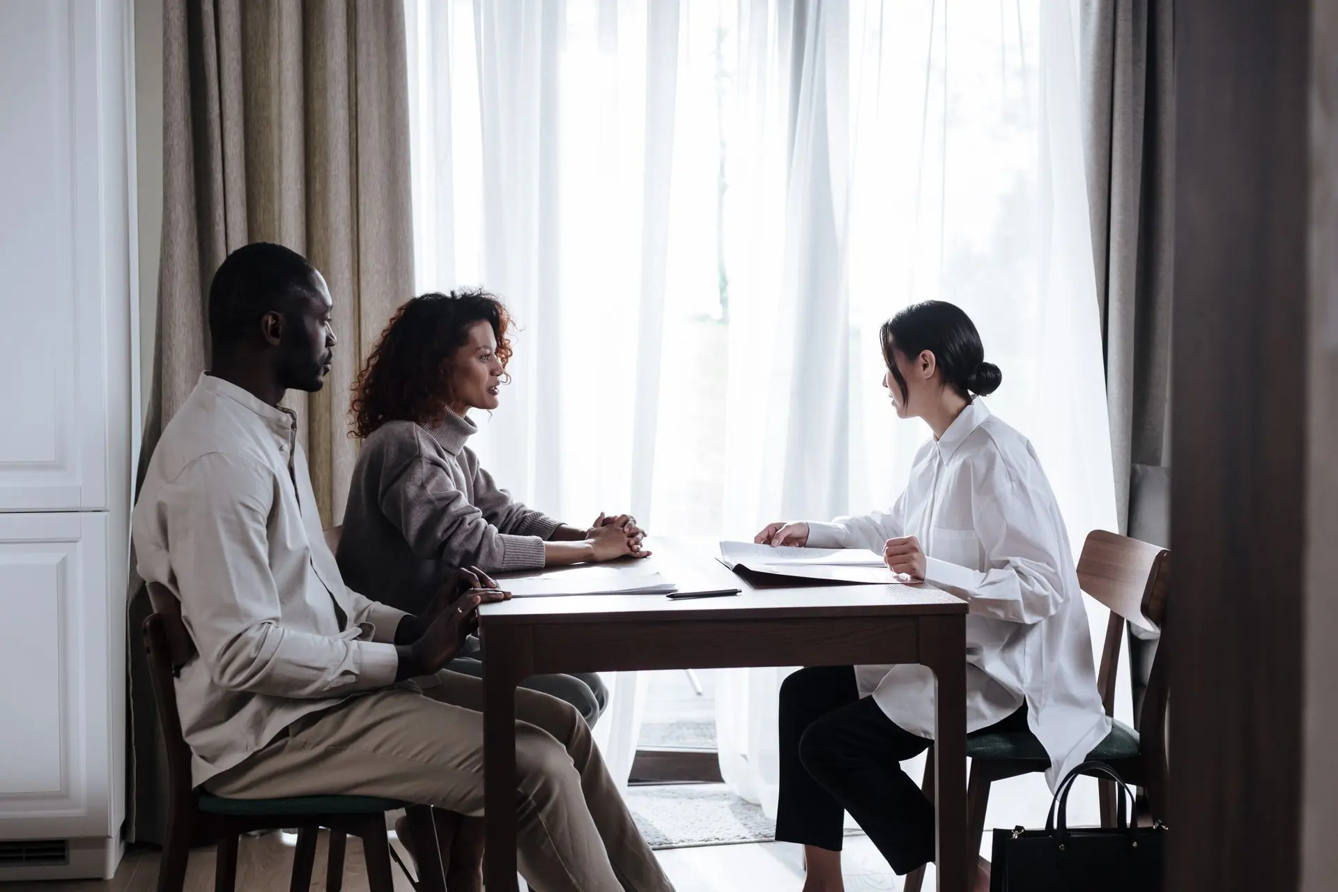 Three people talking across a wooden table with papers in a calm room lit by tall curtained windows