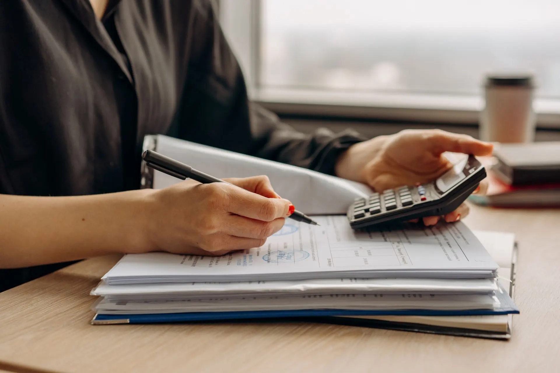 Close-up of hands working through a stack of financial documents with a pen and a calculator beside a window