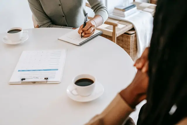 Seen from above: a round white table with two cups of coffee, a printed form on a clipboard and a notebook being written in
