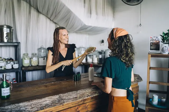 A shop owner behind a wooden counter hands a product to a customer in a bright store lined with jars