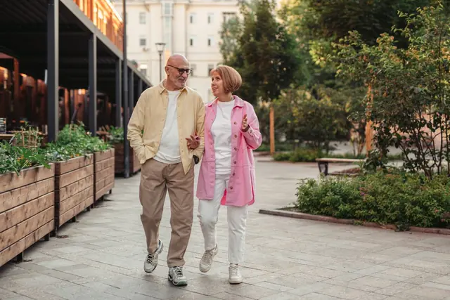 An older couple walking arm in arm along a leafy pavement, chatting and smiling