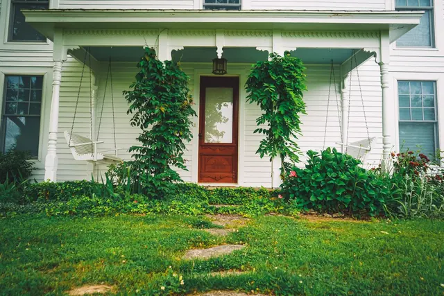 The front porch of a white house with a red door framed by climbing plants and a green lawn