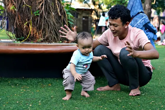 A parent crouches on the grass with open hands as a toddler takes unsteady first steps in a park