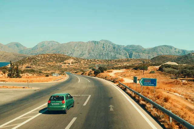 A small green car driving along a winding road through dry hills towards distant mountains