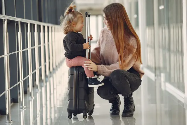 An adult crouches beside a small child sitting on a suitcase in a bright glass-walled airport corridor