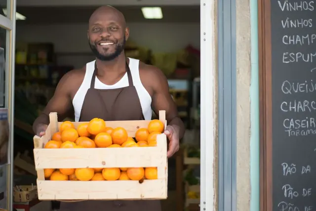 A smiling shopkeeper in an apron carries a crate of oranges out of a small grocery doorway