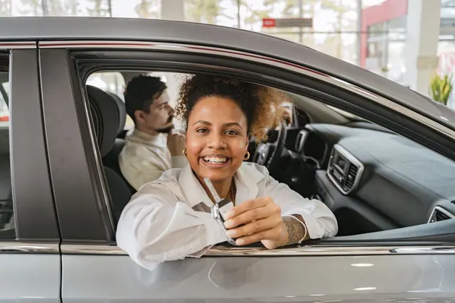 A smiling person leans out of the driver’s window of a new car holding the keys, with a passenger beside them