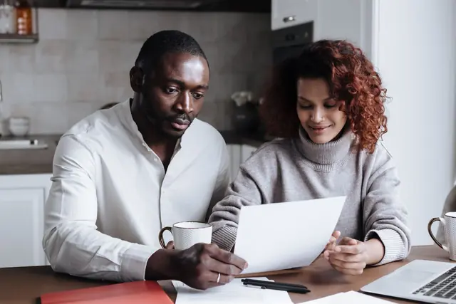 Two people at a kitchen table reading a printed letter together beside a laptop and coffee mugs