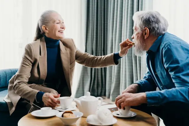 An older couple laughing over tea and biscuits at a small round table in a bright room