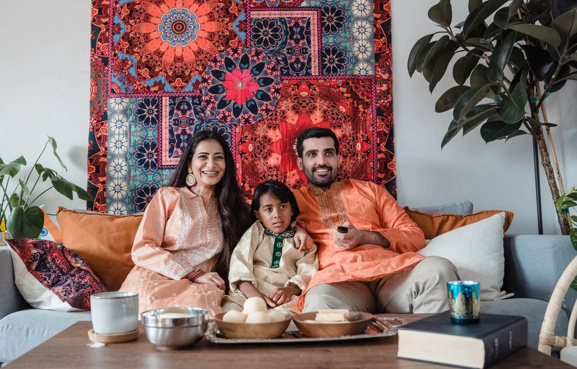 A family of three in festive clothes sitting together on a sofa at home, with snacks on the table in front of them