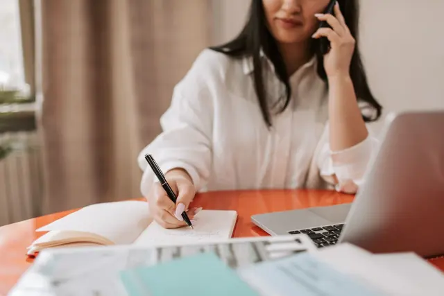 A person on a phone call writes notes in a notebook at a desk beside a laptop